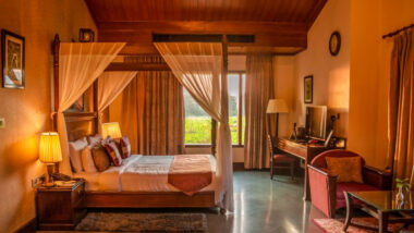 Four poster bed in a room with sofa, table, and an oversized window aahana resort ramnagar