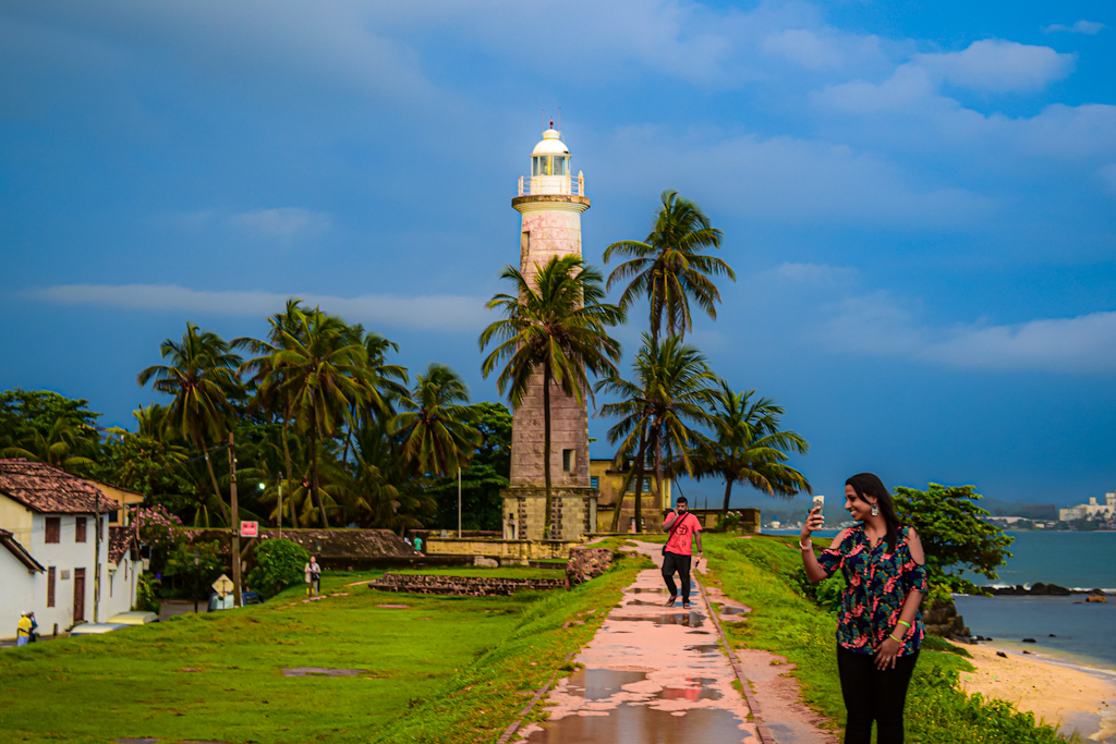 Girl taking selfie with white lighthouse in background and surrounded by sea on one side