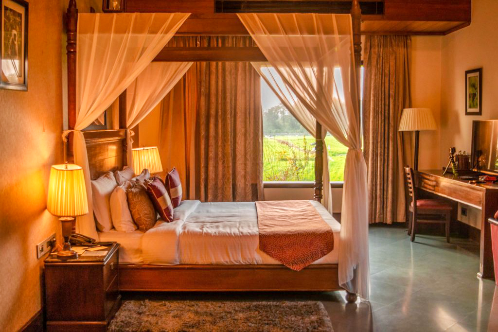 Bed in a room with oversized window looking at the fields aahana Resort ramnagar